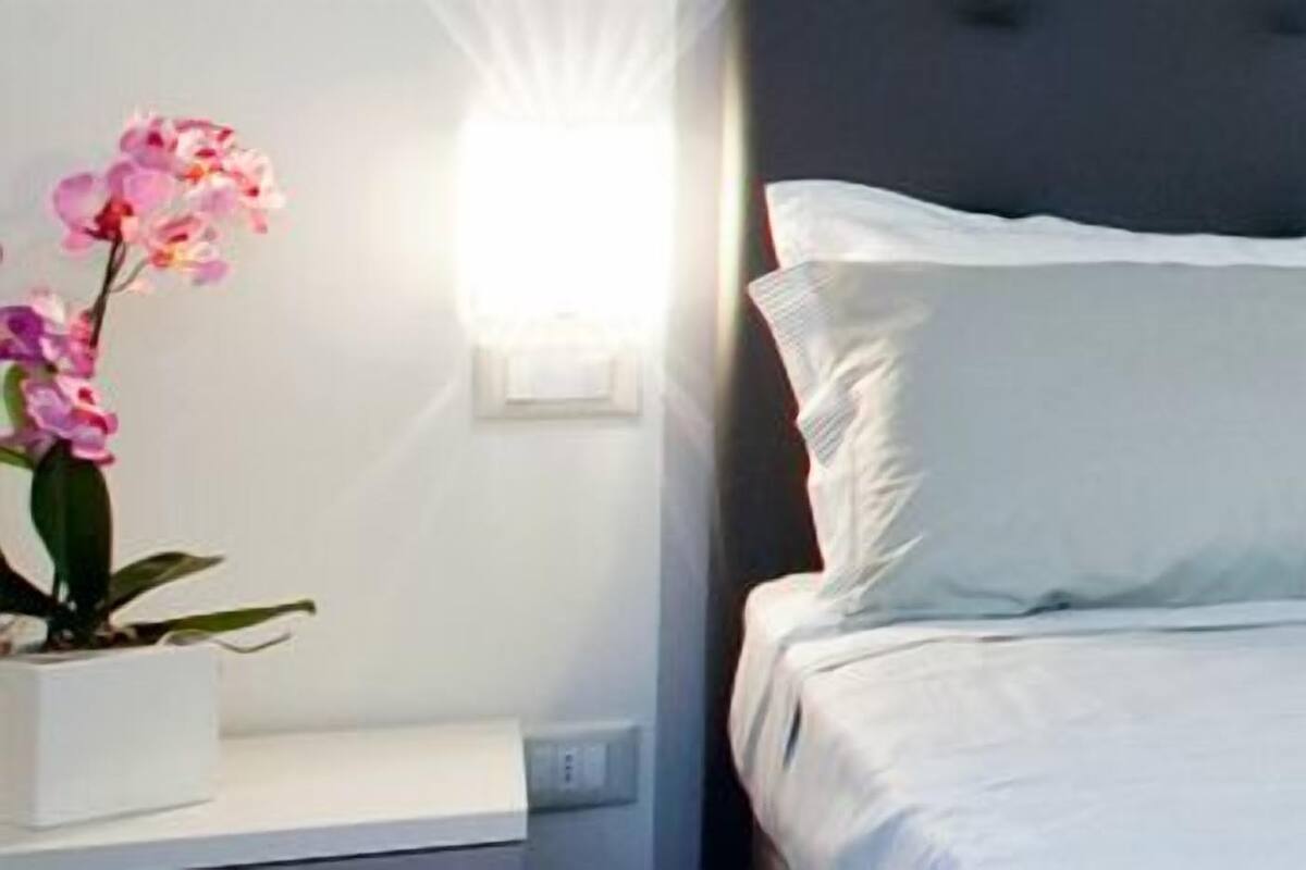 A neatly made bed with white linens and two pillows, a bedside plant, and a wall-mounted light fixture.