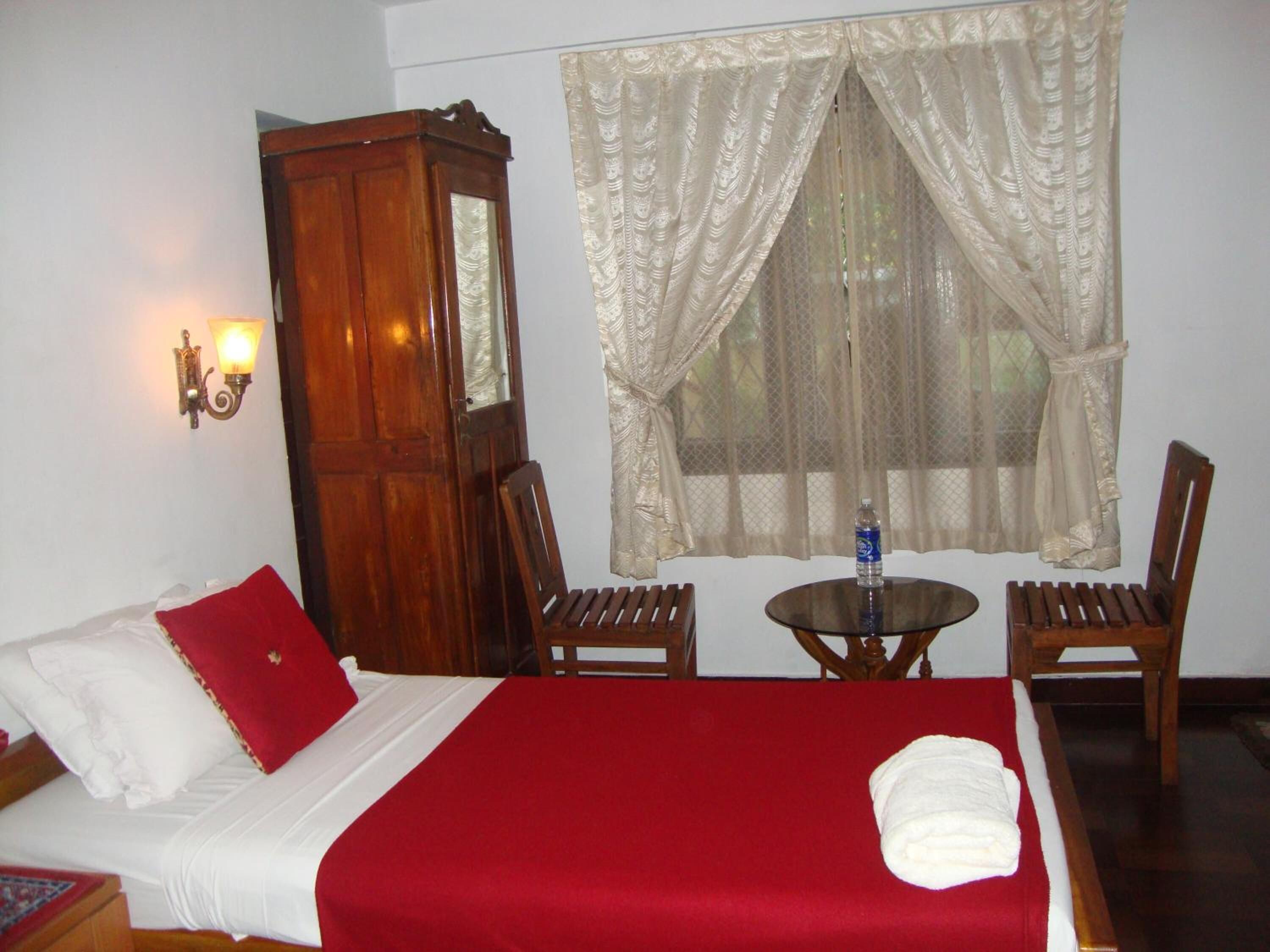 A hotel room with a bed, a wooden wardrobe, a table with chairs, and a window with sheer curtains.