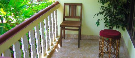 A wooden chair on a balcony with a red cushion, a wicker stool, and a white railing.