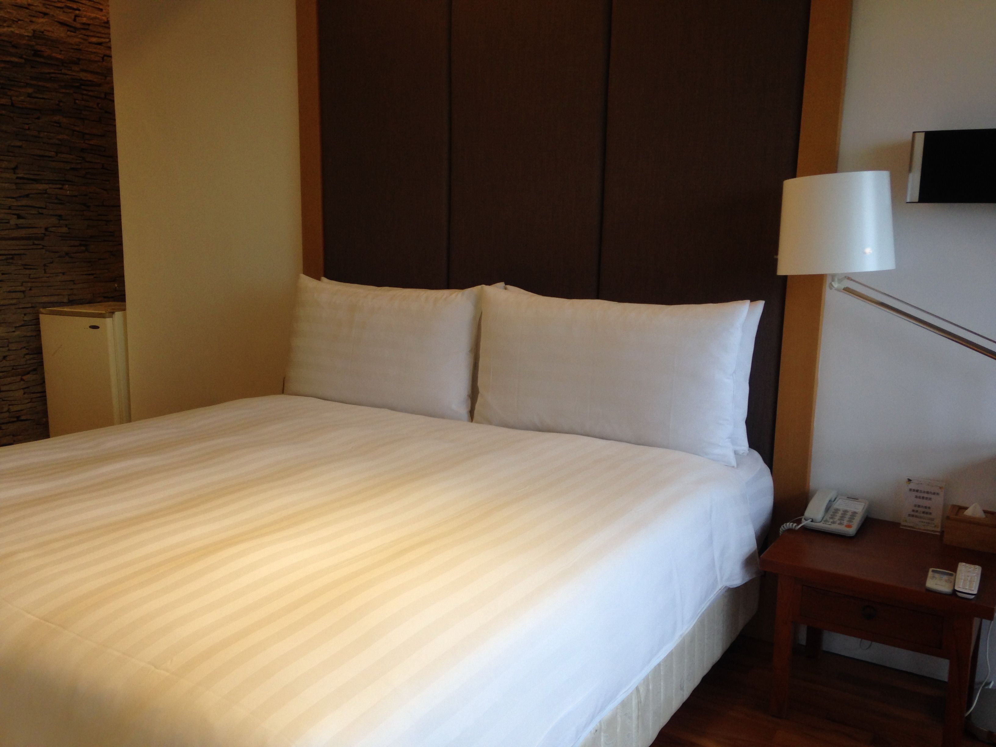 A neatly made bed with white linens, a wooden headboard, a bedside table with a lamp, and a telephone.