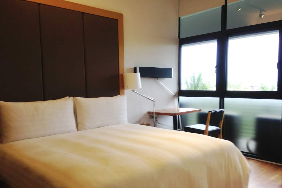 Habitación de hotel con una cama grande, un escritorio y una silla. Se ve follaje verde por la ventana.