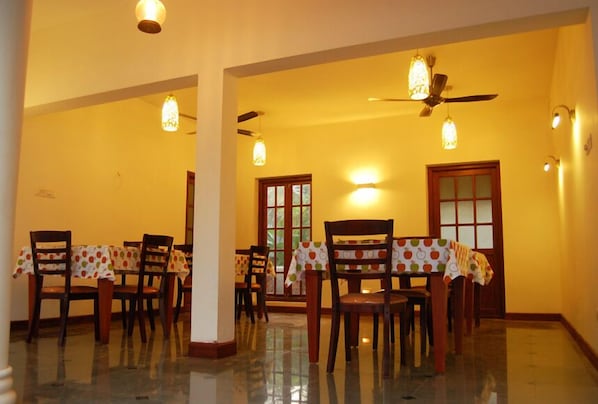 A dining area with a polished floor, wooden chairs, a table with a patterned tablecloth, and ceiling fans. - The Villa Romantica (Aluva)