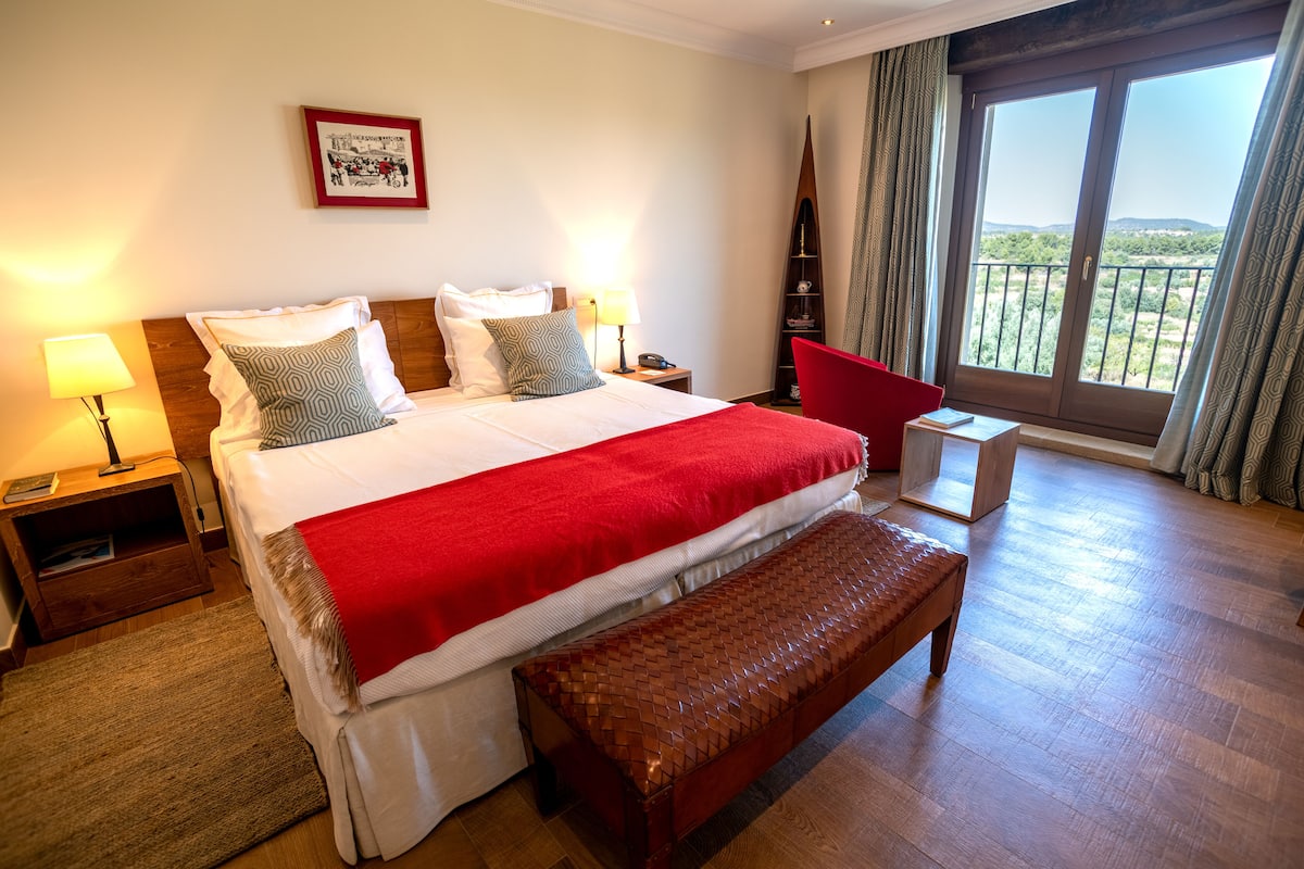 Une chambre d’hôtel avec un grand lit, un banc en bois, une table de chevet avec une lampe et une vue sur l’extérieur grâce à une porte-fenêtre donnant sur un balcon.