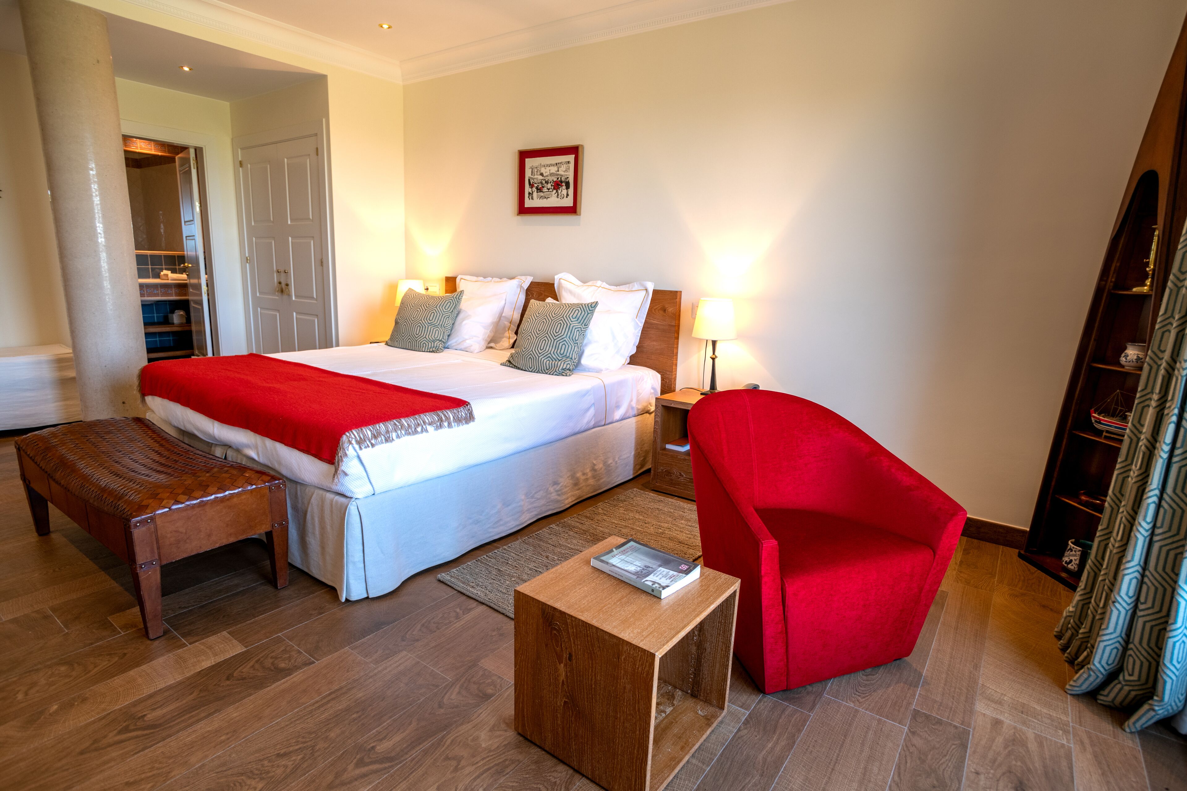 A hotel room with a large bed, a red armchair, a wooden bench, and a small table.