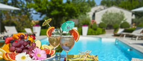 A poolside setting with a fruit platter, two drinks with umbrellas, and a pool in the background.
