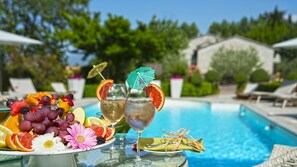 Une scène au bord de la piscine, avec une assiette de fruits, deux boissons décorées de petits parasols et une piscine en arrière-plan.