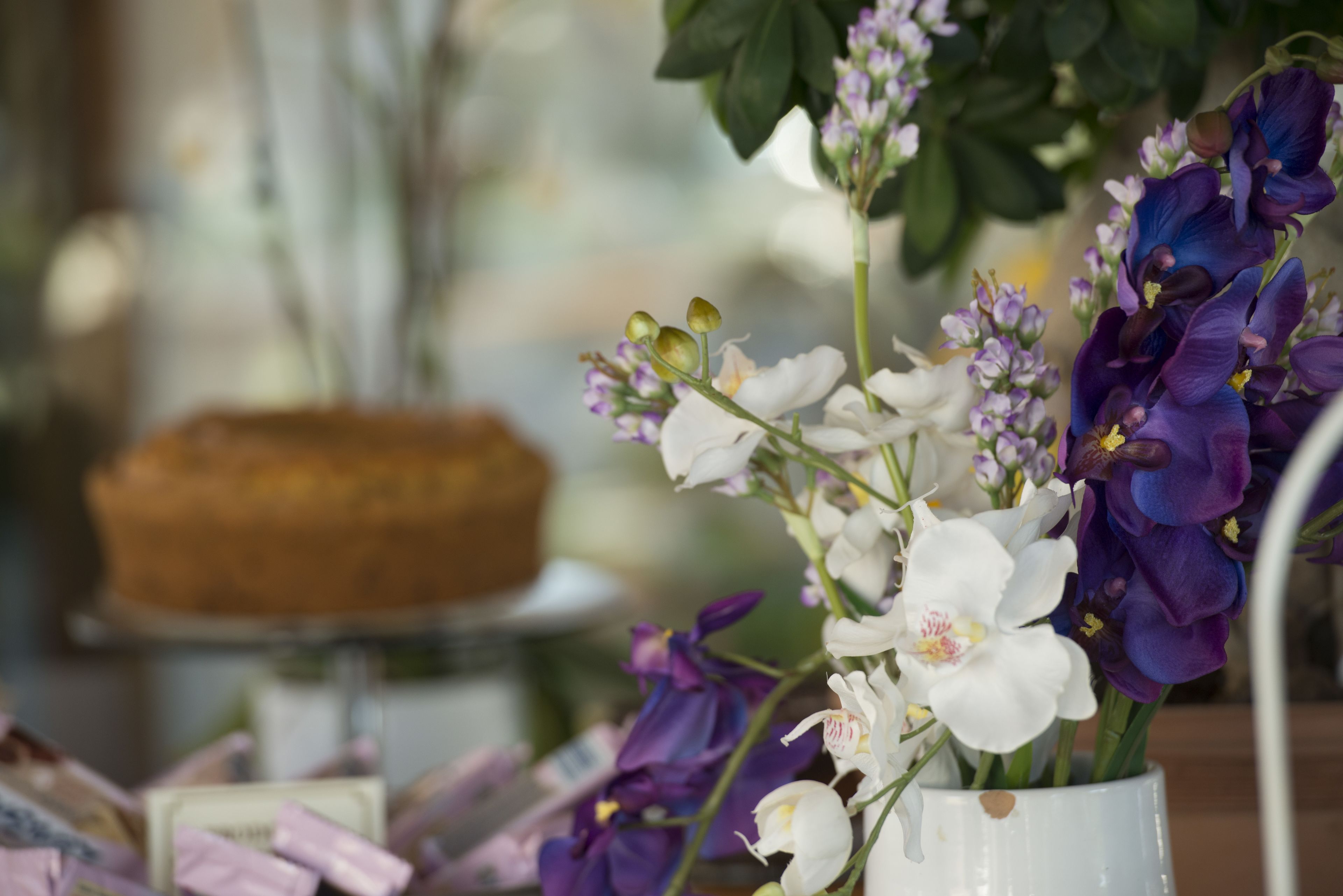 A table with a white vase containing purple and white flowers, and a cake in the background.
