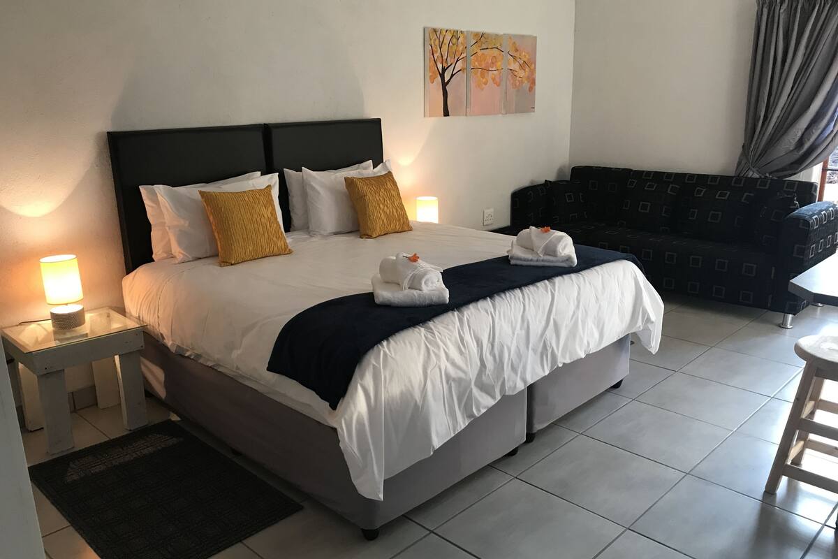 Ein ordentlich bezogenes Bett mit weißen und gelben Kissen, ein dunkles Kopfstück und ein Nachttisch mit Lampe. Es gibt ein dunkles Sofa und ein Bild an der Wand.