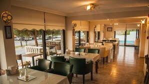 A restaurant with multiple tables and chairs, large windows with a view of parked cars and greenery, and a tiled floor. - Sun Hotel (Cesme)