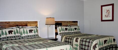 Ein Zimmer mit zwei Betten, jedes mit einem zudeckenden Waldmotiv. Zwischen den Betten steht ein Nachttisch mit einer Lampe.