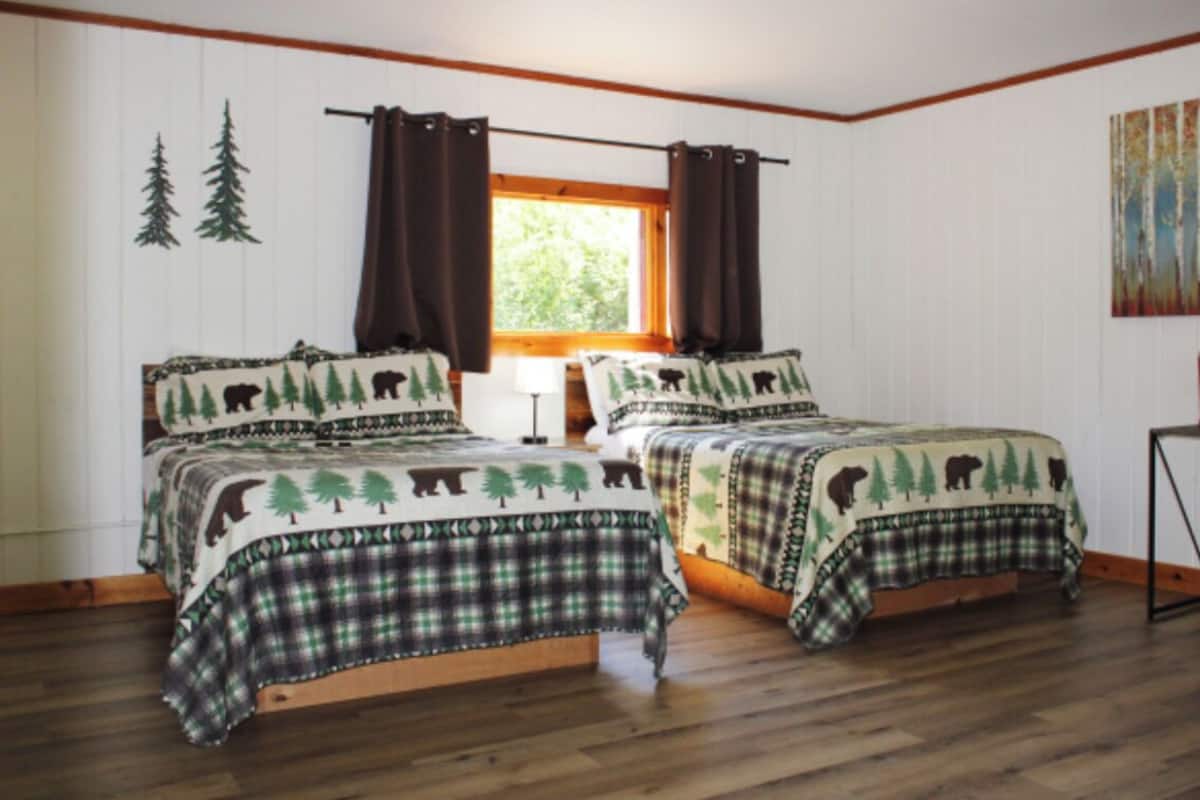 A room with two beds, each with a forest-themed bedspread, a window with curtains, a painting on the wall, and a small table with a red object on it.