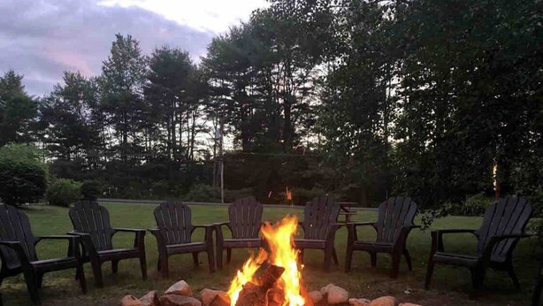 A fire pit with logs surrounded by rocks, and several Adirondack chairs arranged around it. - Luzerne Court Motel (Lake Luzerne)
