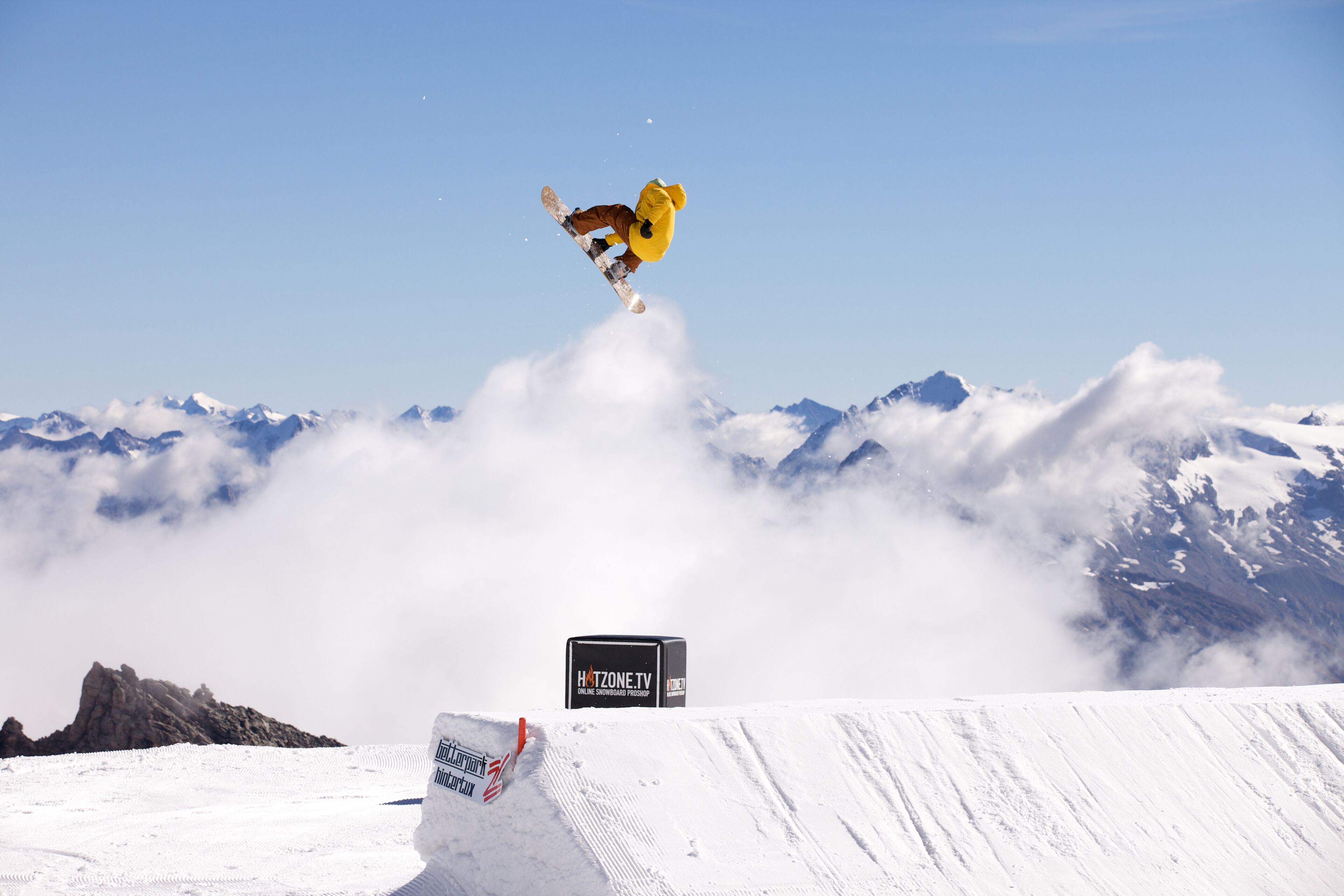 Un snowboarder realizando un salto sobre una rampa de nieve, con montañas al fondo.