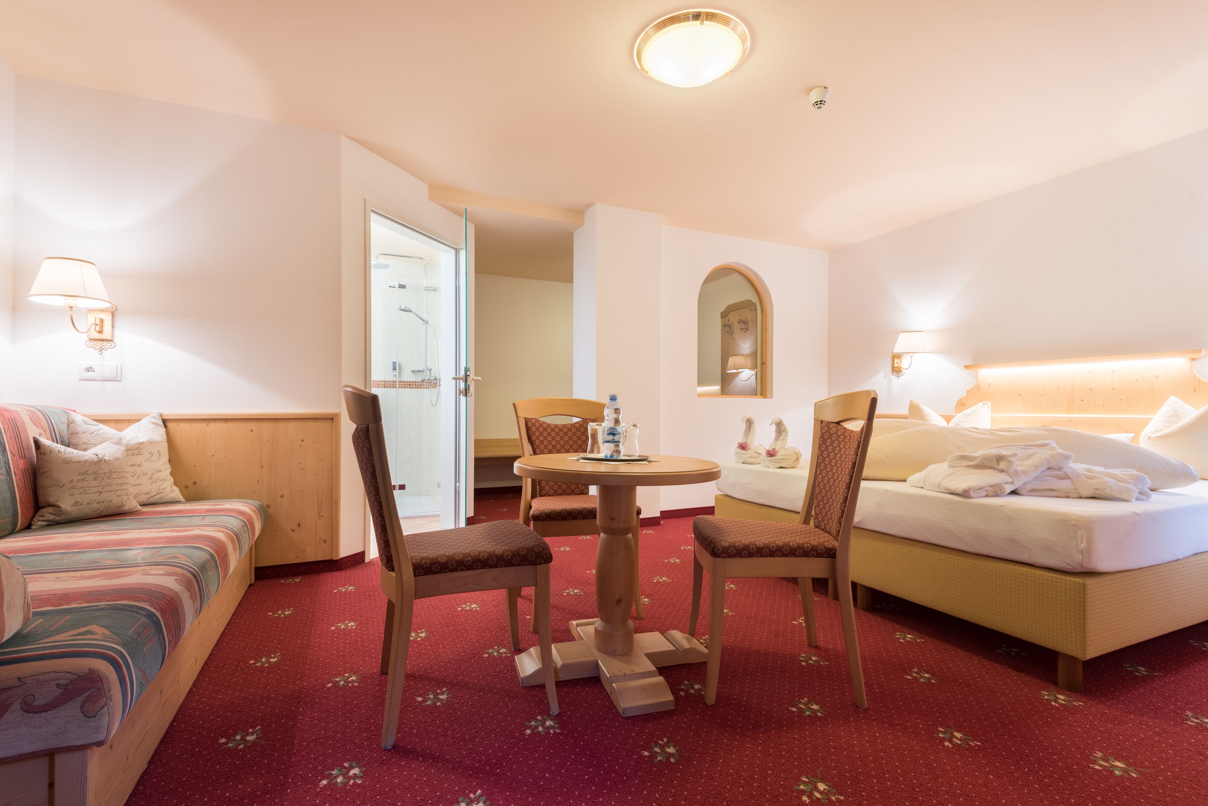 Habitación de hotel con cama, sofá, mesa de comedor con sillas y baño visible a través de una puerta abierta.