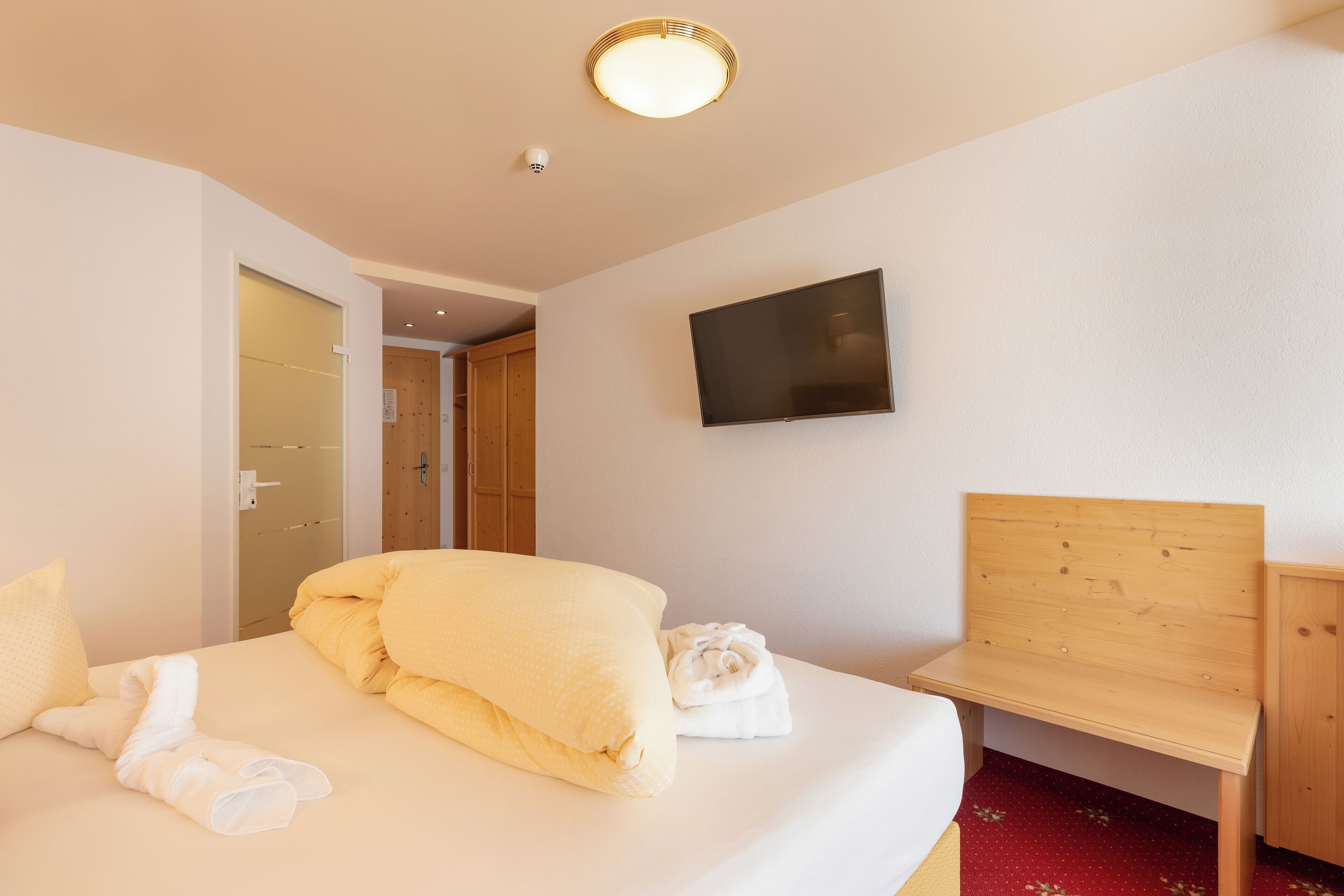 A hotel room with a bed, a wooden bench, a TV mounted on the wall, and a door leading to another room.