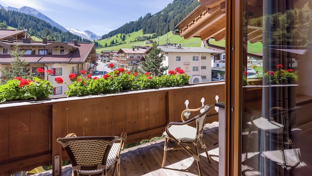 A balcony with wicker chairs, a wooden railing, and a view of a mountain town with red flowers in the foreground. - Hotel Pinzger (Tux)