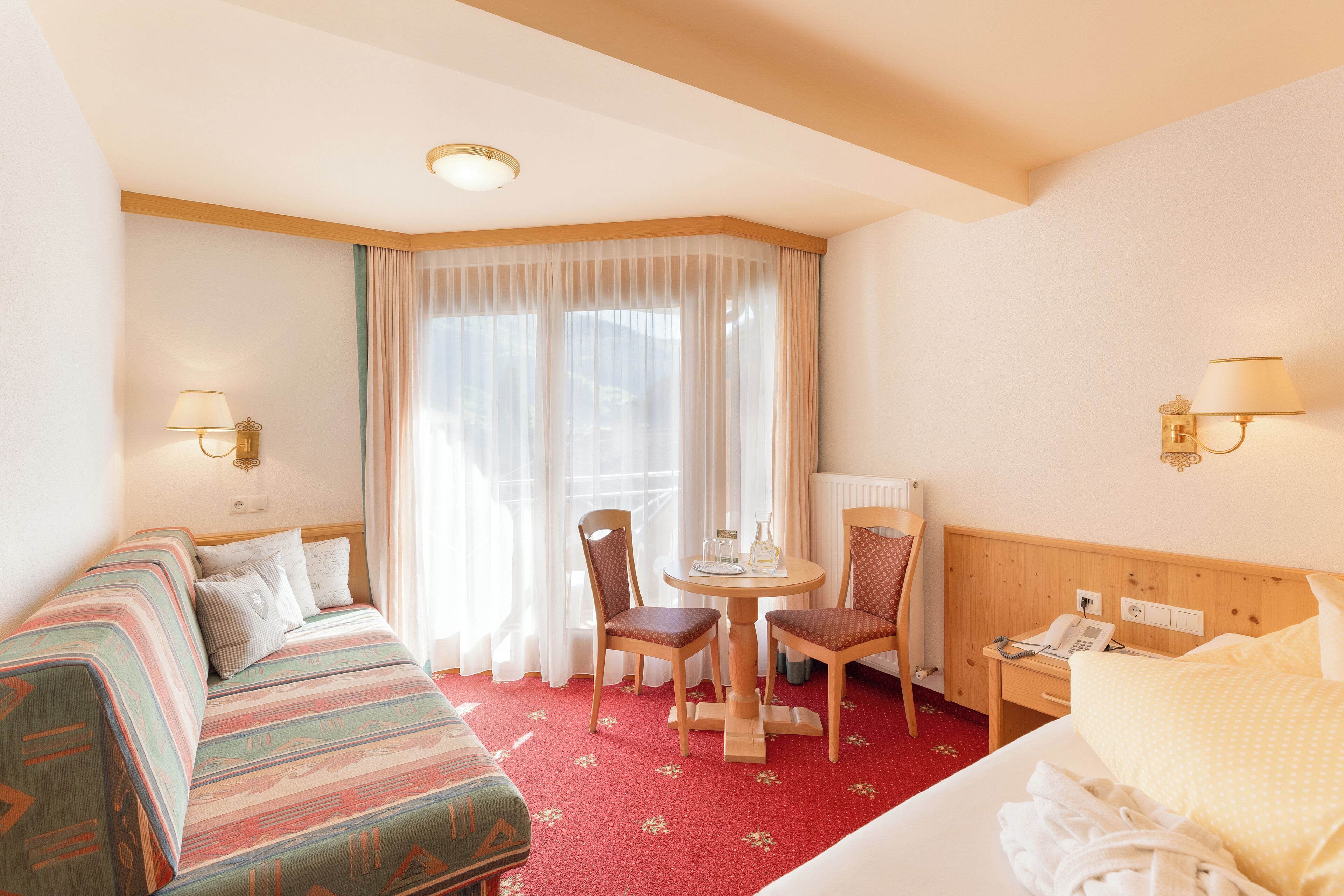 A hotel room with a bed, a sofa, a dining table with chairs, a small table with a lamp, and a telephone.