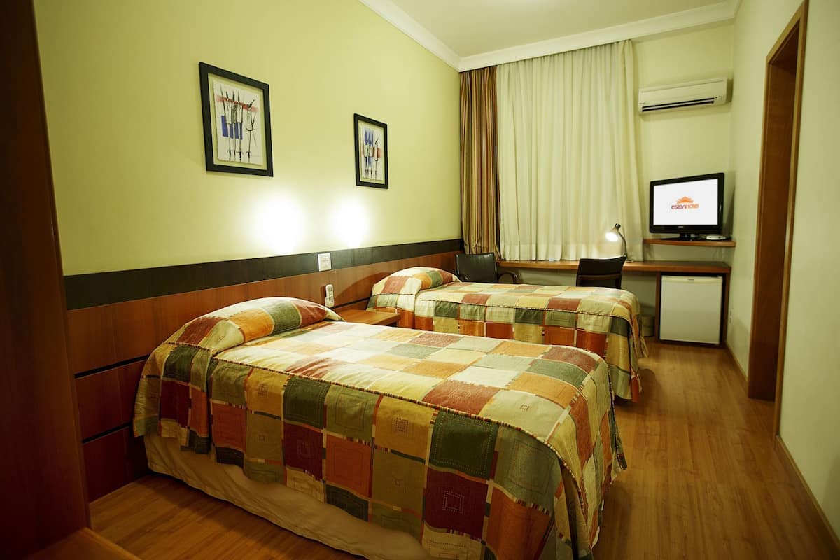 A hotel room with two beds, a desk with a computer, a television, and an air conditioning unit.