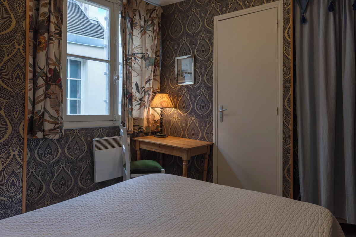 A bedroom with a bed, a desk with a lamp, a radiator, and patterned wallpaper.