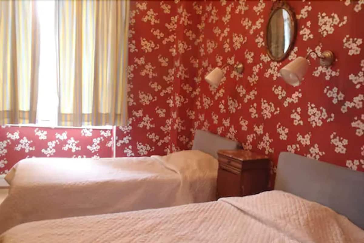 A room with two beds, a small wooden nightstand, a mirror on the wall, and a window with curtains.
