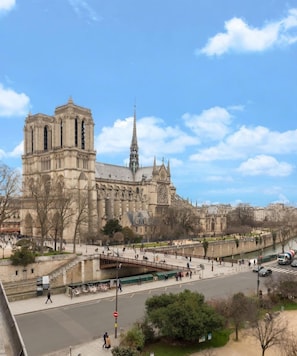 Una catedral histórica con una torre del reloj destacada, un río en primer plano y un puente peatonal.
