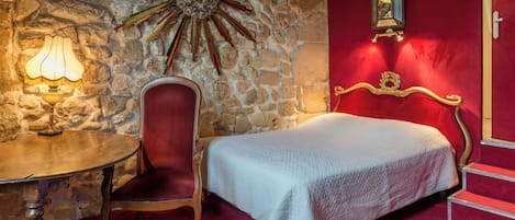 Una habitación con pared de piedra, pared roja, cama con colcha blanca, una silla, una mesa con lámpara y un espejo adosado a la pared.