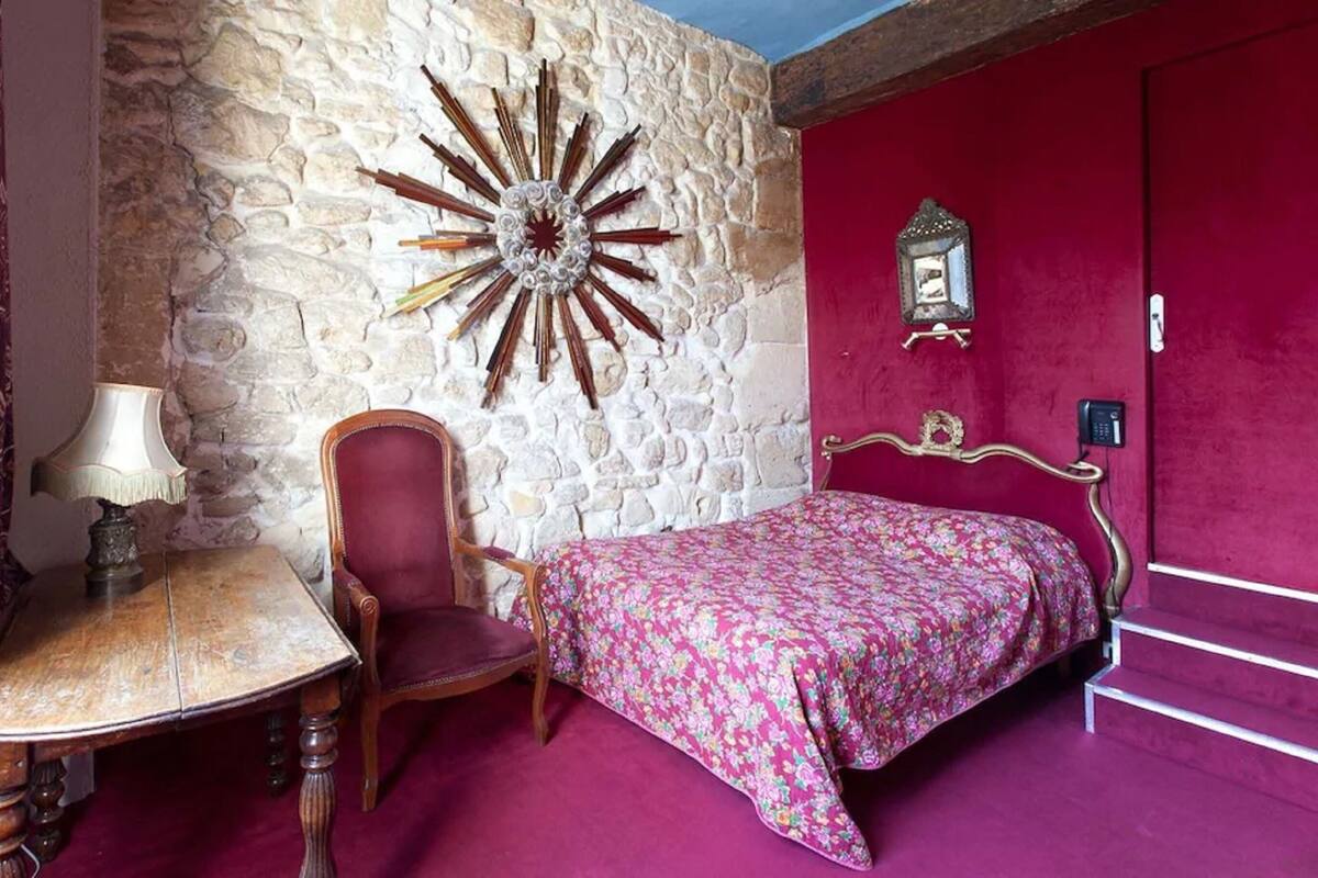 A bedroom with a stone wall, a bed with a floral bedspread, a red chair, a wooden table with a lamp, and a red door.
