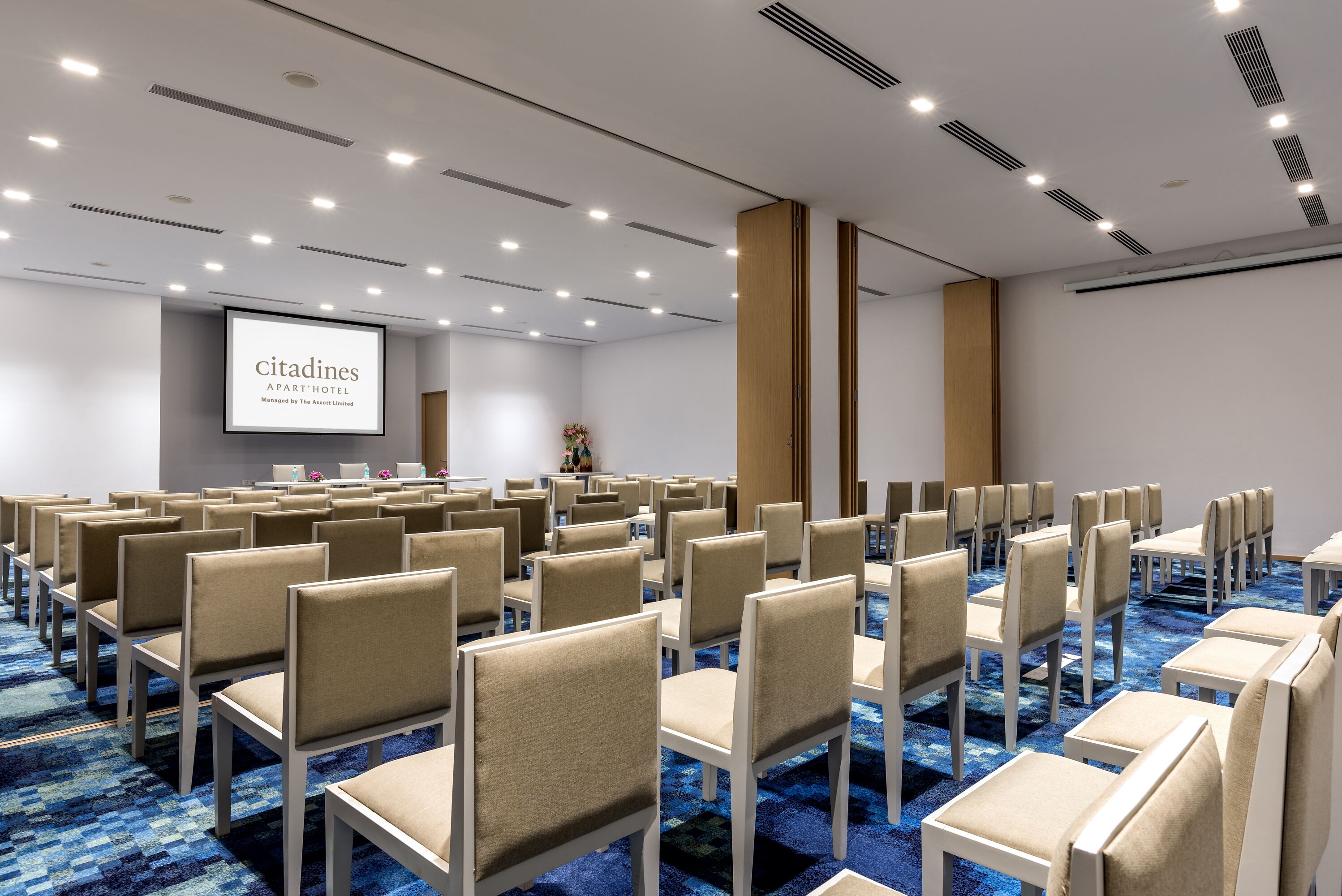 A conference room with rows of beige chairs, a large screen displaying 'citadines', and a blue carpeted floor.