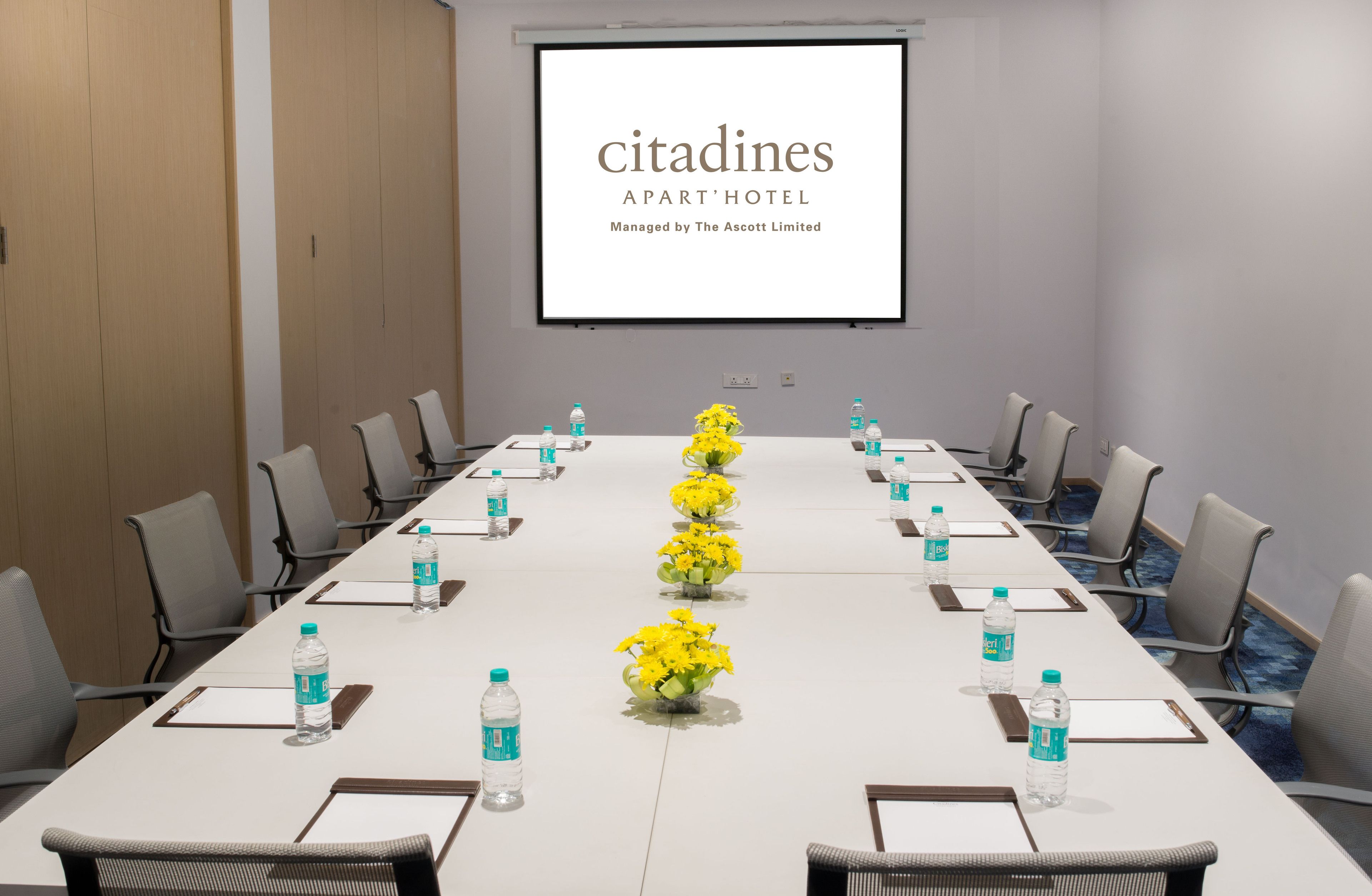 A conference room with a long table, chairs, water bottles, and flower centerpieces. A screen displays the text "citadines APART' HOTEL Managed by The Ascott Limited".