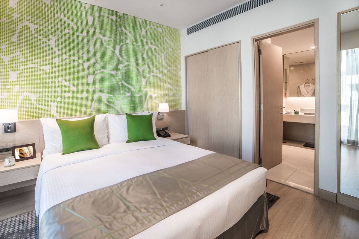 Ein modernes Hotelzimmer mit einem großen Bett, einem Nachttisch, einer Wand mit grünem Muster und einem durch eine offene Tür einsehbaren Badezimmer.