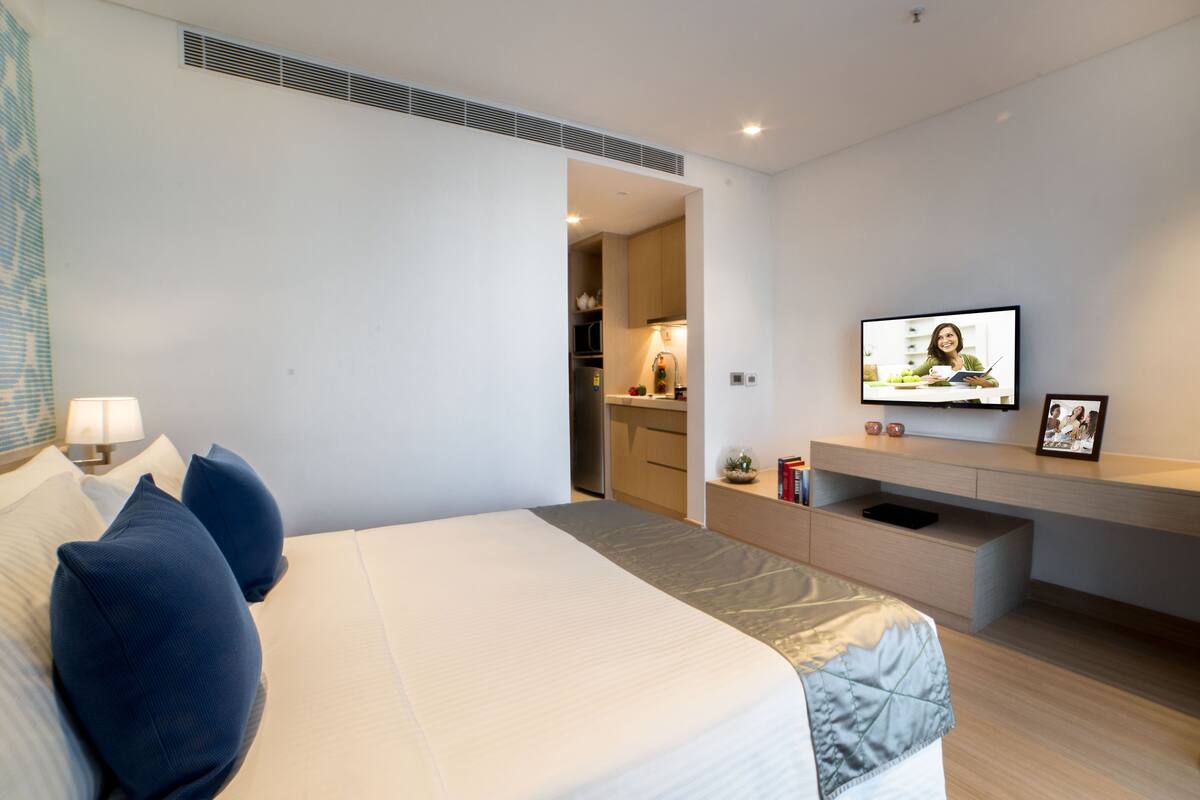 Ein modernes Hotelzimmer mit einem großen Bett, einem an der Wand montierten Fernseher und einer kleinen Küchenzeile im Hintergrund.