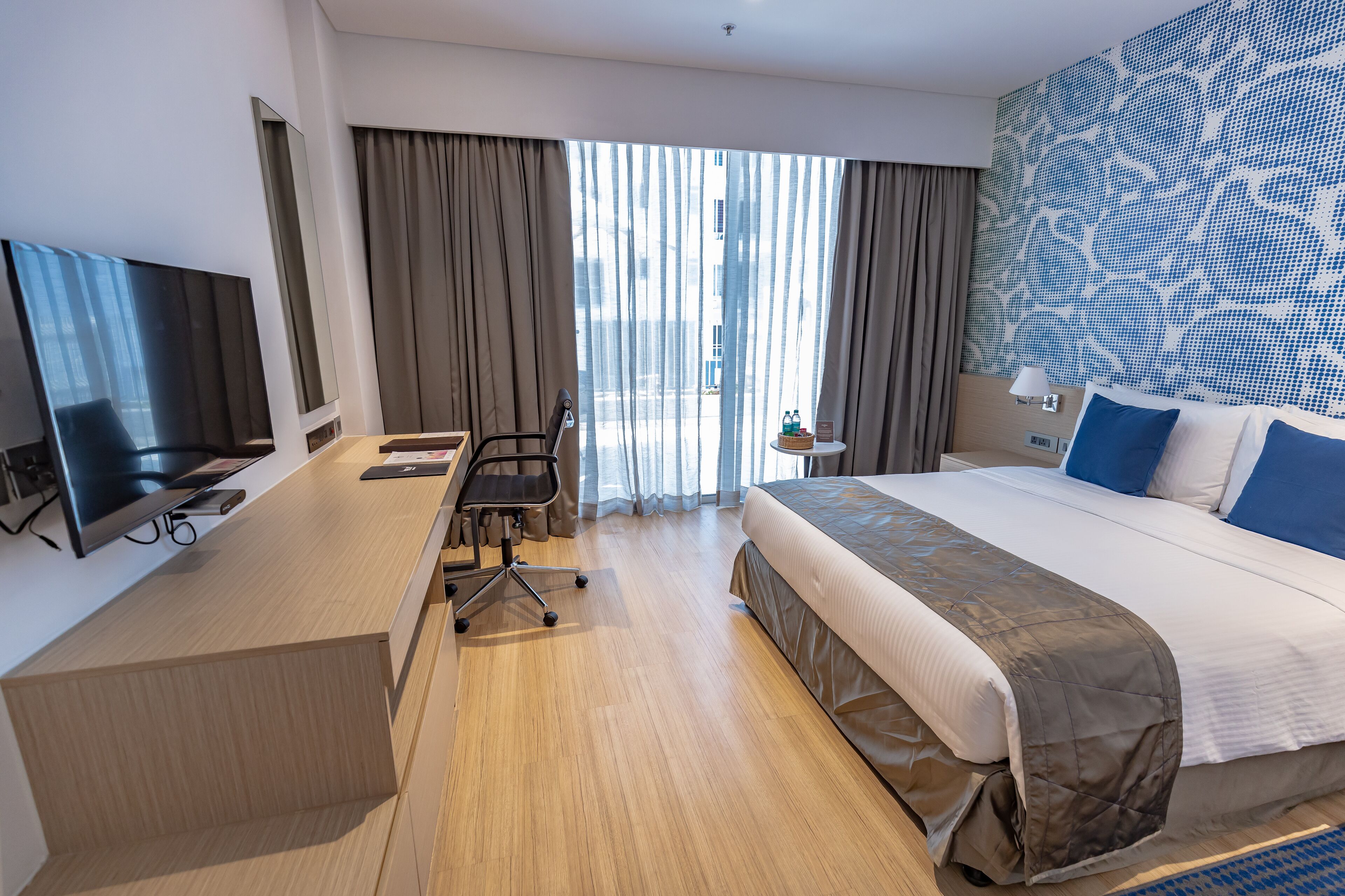 A modern hotel room with a large bed, a desk, a flat-screen TV, and a view of the city through large windows.