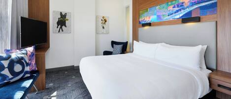 A hotel room with a large bed, a TV mounted on the wall, a chair with a patterned cushion, and artwork on the walls.