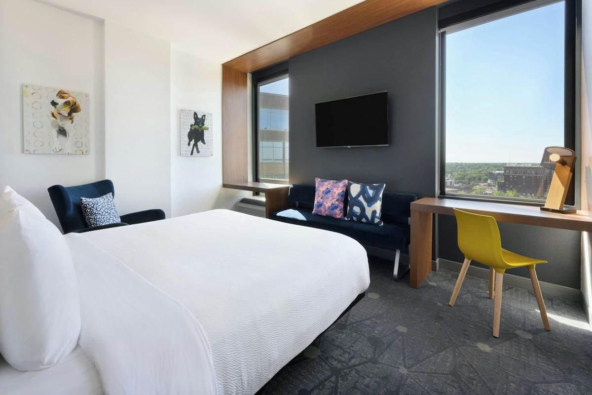 Habitación de hotel con una cama grande, un escritorio con silla, un televisor y una ventana con vistas a la ciudad.