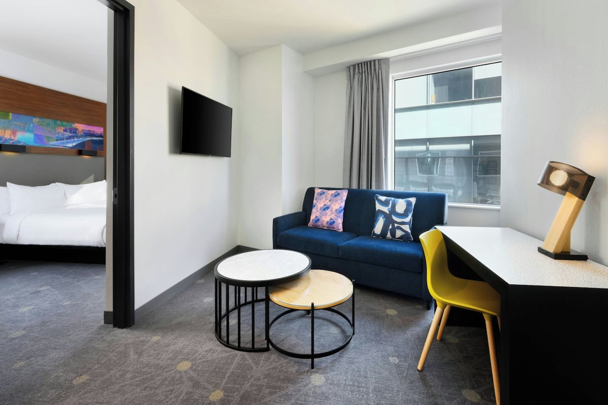 Habitación de hotel con cama, sofá, escritorio, silla, televisor y ventana con cortinas.