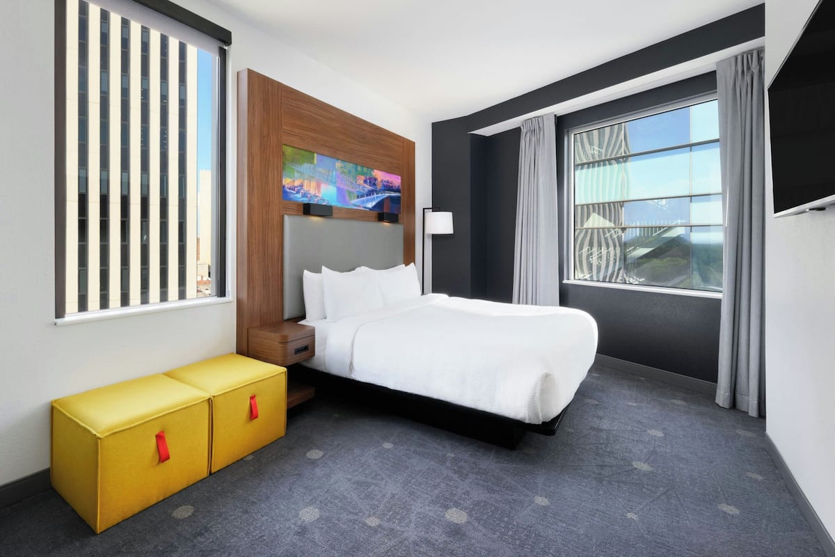 Una habitación de hotel moderna con una cama grande, un cabecero de madera, un cuadro colorido, una mesita de noche y una ventana con cortinas.