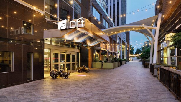 L’entrée d’un hôtel au design moderne, avec une enseigne « aloft » bien visible, une galerie couverte et un éclairage décoratif.