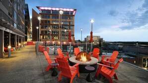 Une terrasse sur le toit avec un foyer entouré de chaises rouges, un bâtiment moderne en arrière-plan et un ciel dégagé.