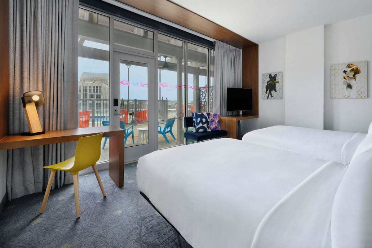 Habitación de hotel con una cama grande, un escritorio con silla, un televisor y un balcón con sillas de colores.