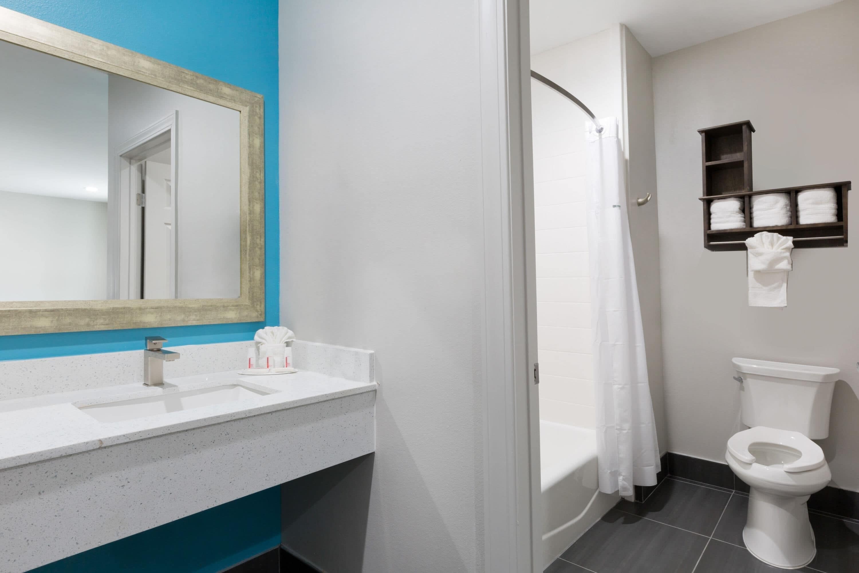A bathroom with a white sink, a mirror, a shower curtain, a toilet, and a shelf with folded towels.