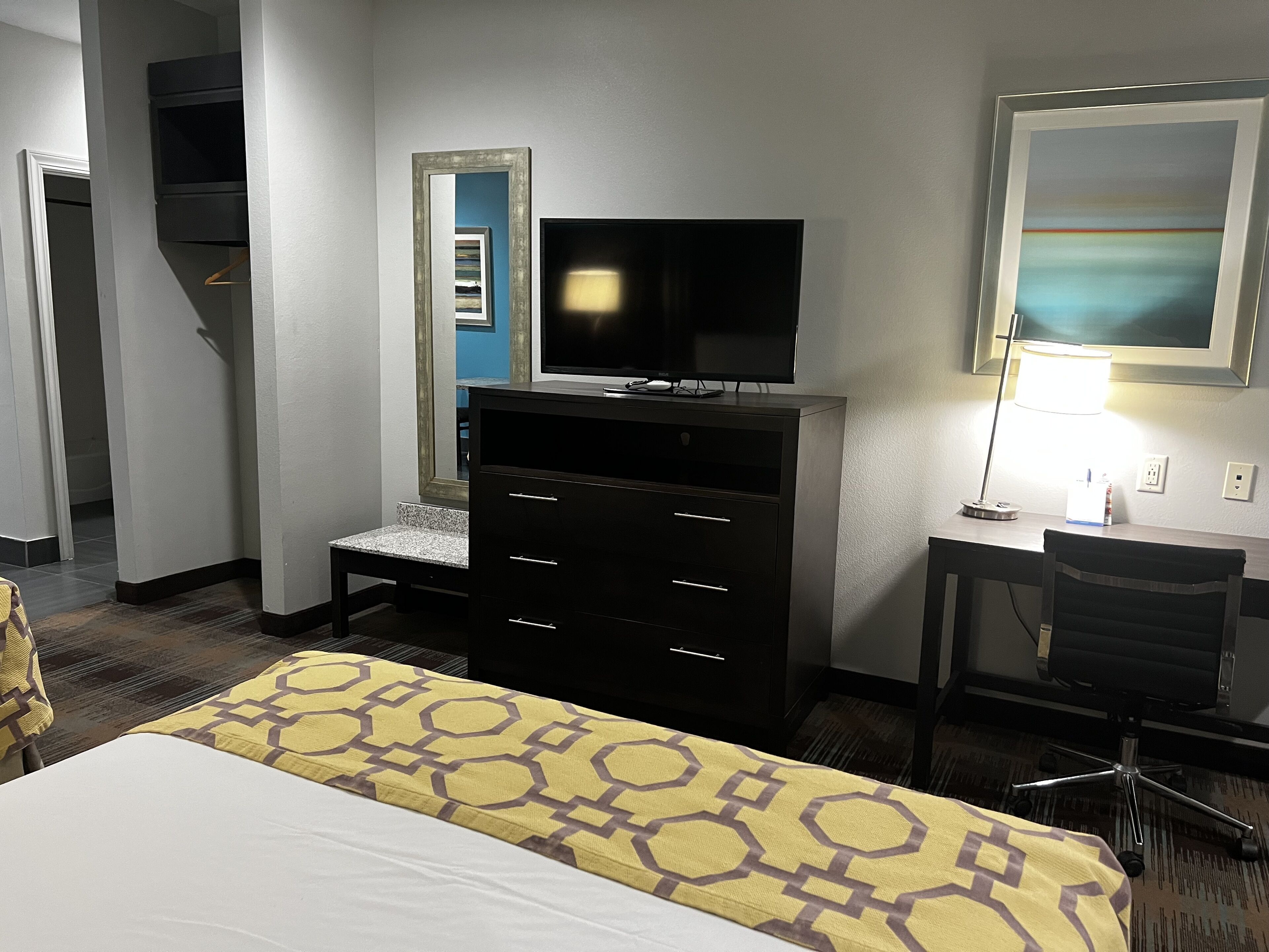 A hotel room with a bed, a desk with a chair, a television, a dresser, and a lamp.