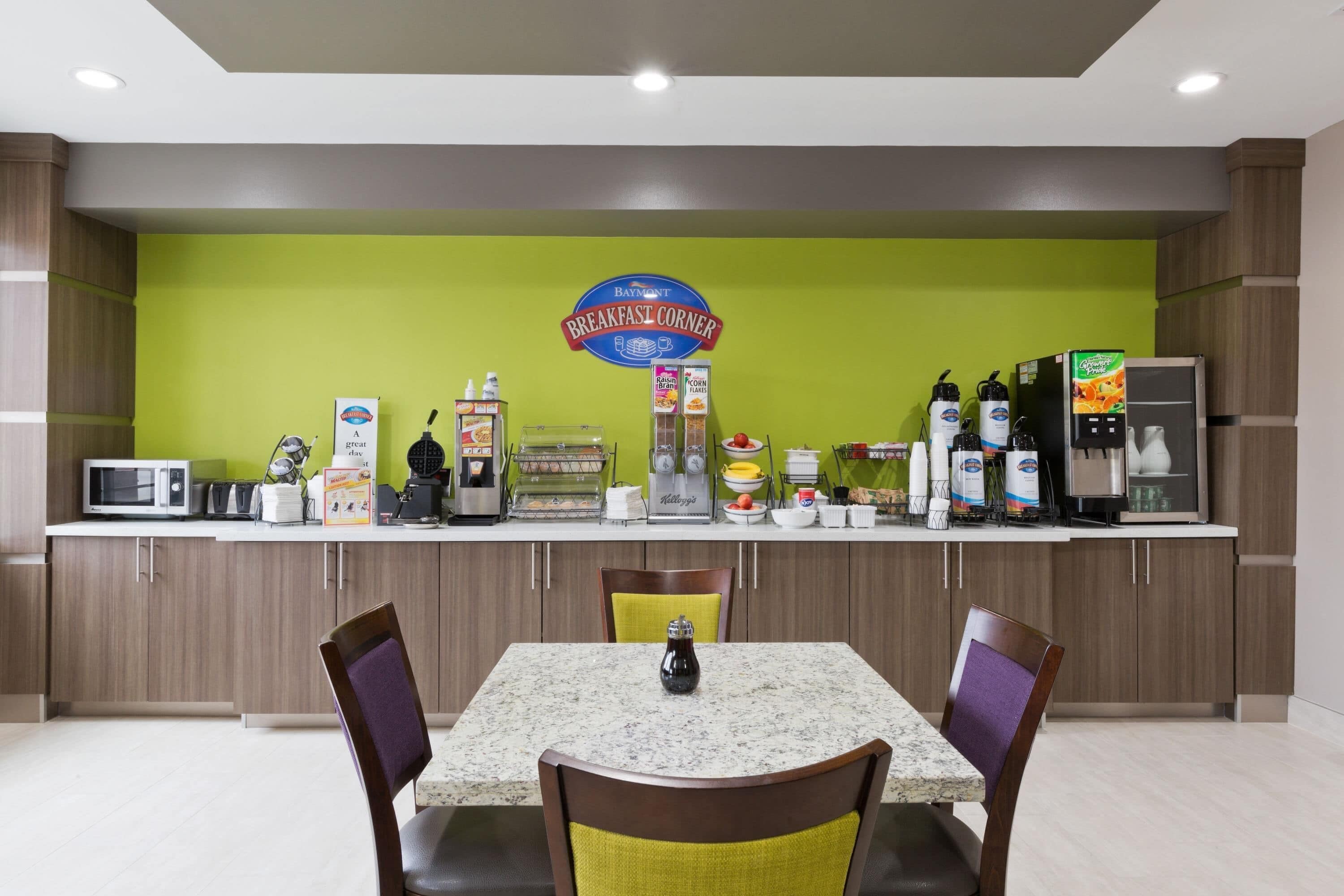 A breakfast counter with a sign reading 'Baymont Breakfast Corner,' equipped with a coffee machine, microwave, and various food items.