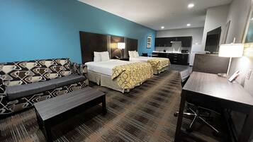 Habitación de hotel con una pared azul de acento, un sofá, dos camas con cubrecamas estampados, un escritorio y una zona de kitchenette.