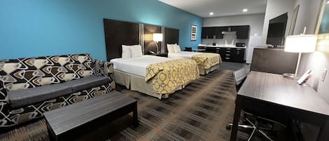 Una camera d'albergo con una parete azzurra, un divano, due letti con coperte a motivi decorativi, una scrivania e un angolo cottura.