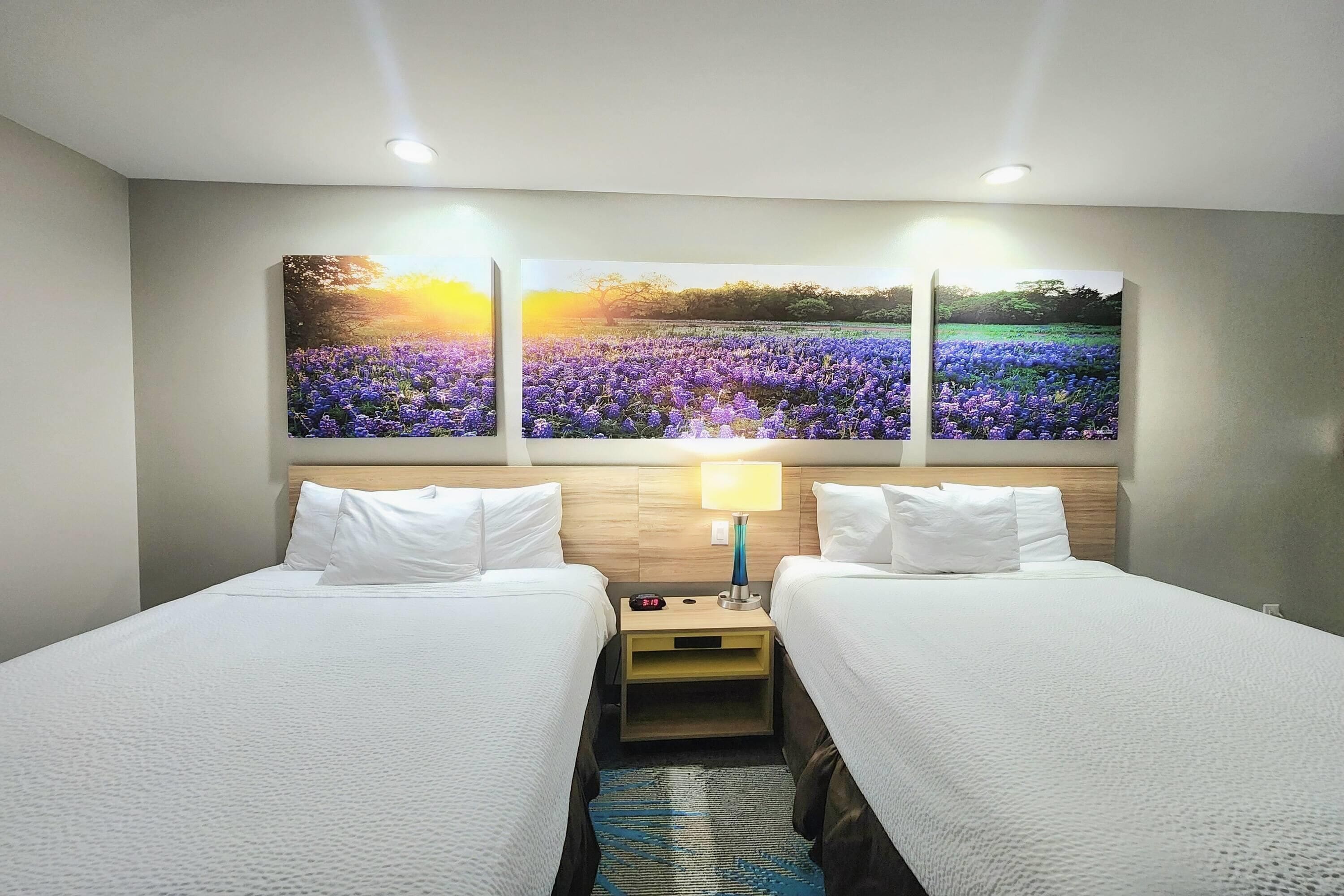 Une chambre d’hôtel avec deux lits, une table de chevet avec une lampe et trois grandes peintures accrochées au mur représentant un champ au coucher du soleil.