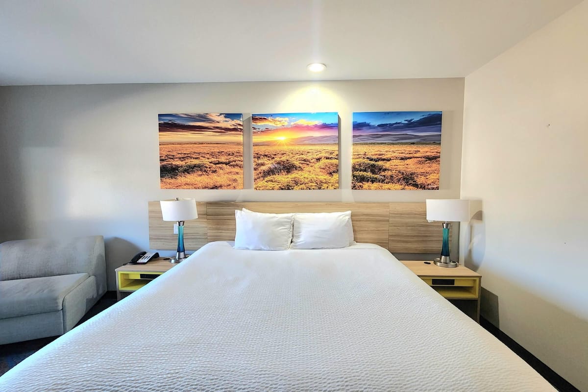 Habitación de hotel con una cama grande, dos mesitas de noche con lámparas, un sofá y un cuadro colgado en la pared que representa un paisaje al atardecer.