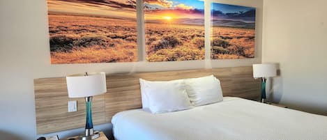 Habitación de hotel con una cama, dos lámparas de noche, una mesita de noche con un teléfono y tres cuadros con paisajes en la pared.