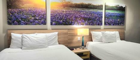 Una habitación de hotel con dos camas, un cabecero de madera, una mesita de noche con una lámpara y un cuadro floral grande sobre las camas.