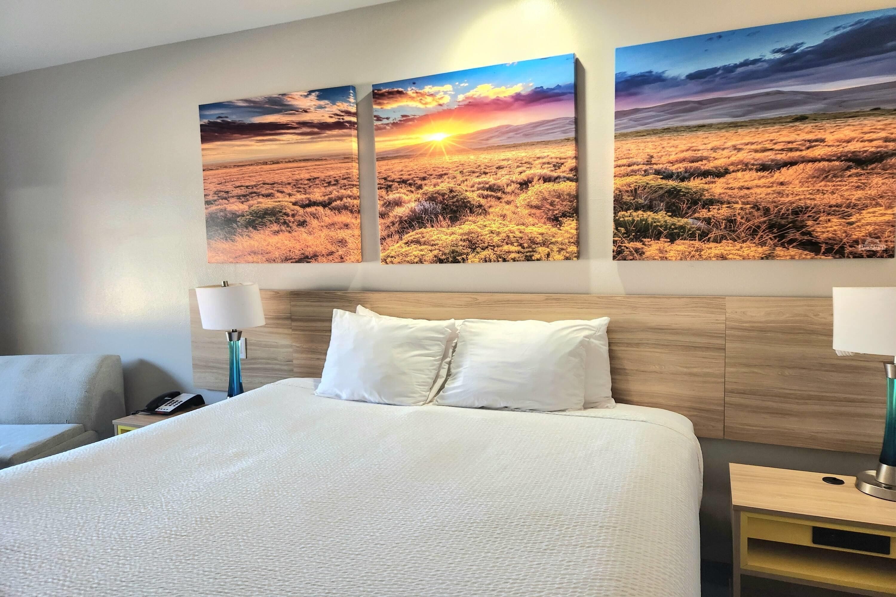 Habitación de hotel con una cama grande, dos mesitas de noche y una obra de arte colgada en la pared que muestra un atardecer sobre un campo.