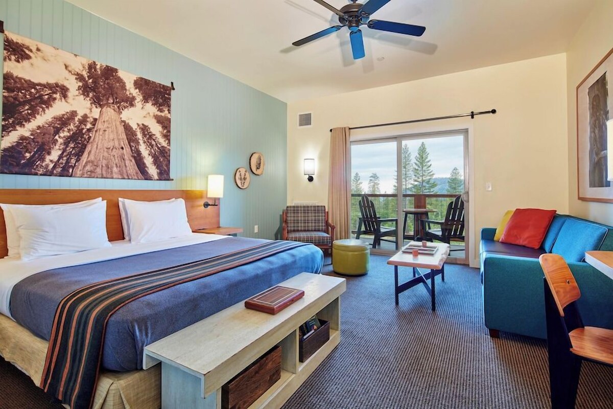 A hotel room with a bed, a desk, a chair, a sofa, a ceiling fan, and a large window.